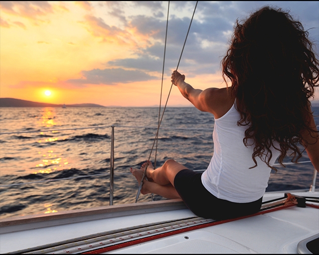 Femme de dos assise sur le bord d'un voilier face au soleil couchant